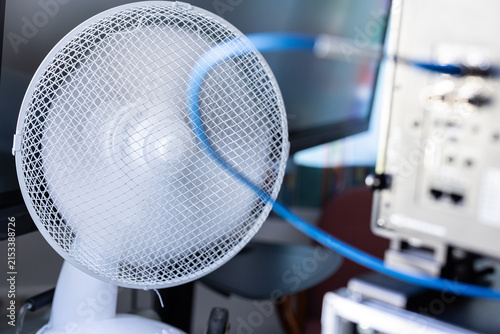 A close-up shot of a white desk fan blowing air near computer monitors and electronic server equipment, with a blurred blue cable running past the foreground.
