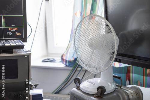 A close-up shot of a white desk fan blowing air near computer monitors and electronic server equipment, with a blurred blue cable running past the foreground.