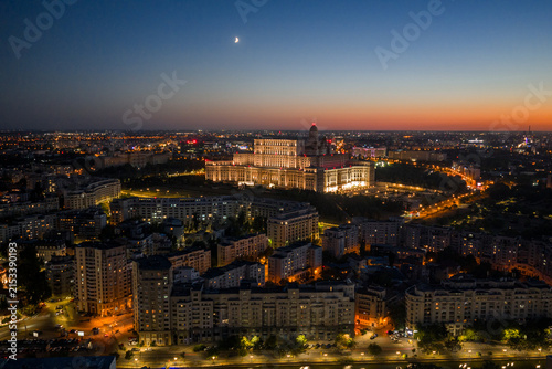 Aerial panoramic night view of the Palace of the Parliament in Bucharest, Romania, illuminated under a twilight sky with a crescent moon and vibrant glowing city lights at sunset.