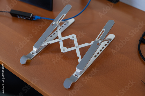 An adjustable, silver aluminum laptop stand set up on a wooden desk surface, featuring anti-slip rubber pads and a foldable scissor mechanism in an office environment.