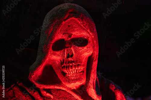 Scary grim reaper skull statue in a hooded cloak illuminated by eerie red light on black background copy space