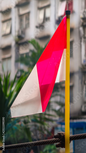 A miniature Indonesian flag. A small red-and-white plastic flag attached to a yellow straw against a backdrop of apartment buildings, in celebration of Indonesia's Independence Day on August 17th.