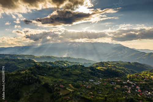 Scenic aerial view of the Rucar-Bran corridor in Romania with dramatic sunbeams breaking through clouds over lush green rolling hills and Carpathian mountains at sunset.