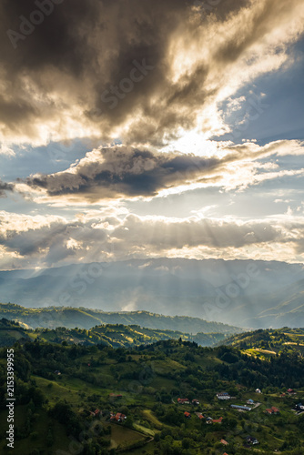 Majestic sunset view over rolling green hills and village in Sirnea, Rucar region, Romania, featuring dramatic cloud formations with golden sunbeams breaking through sky.
