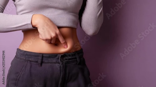 A young woman with long dark hair points to her stomach. She wears a light gray long-sleeve top and dark pants. The background is a soft purple color.