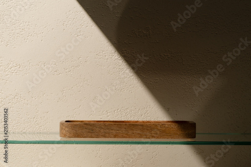Small empty wooden tray placed on a transparent glass shelf in front of a travertine effect plaster wall with a dramatic diagonal shadow, serving as a minimal cosmetic product staging prop.