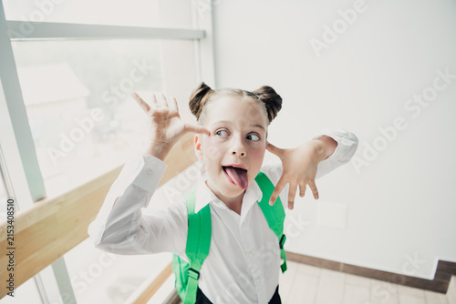 Obraz na plátne Silly school girl making a funny face with tongue out and green backpack before