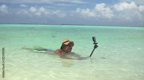 Man floating face down in shallow tropical beach water while taking a selfie with an action camera mounted on a selfie stick, capturing travel vlog, vacation, and outdoor recreation.