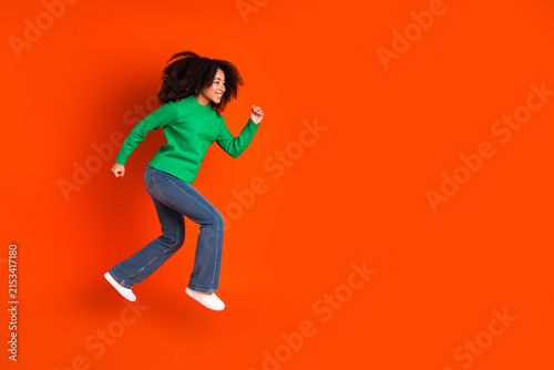 Canvastavla Young woman jumping in green sweater and jeans against a vibrant orange backgrou