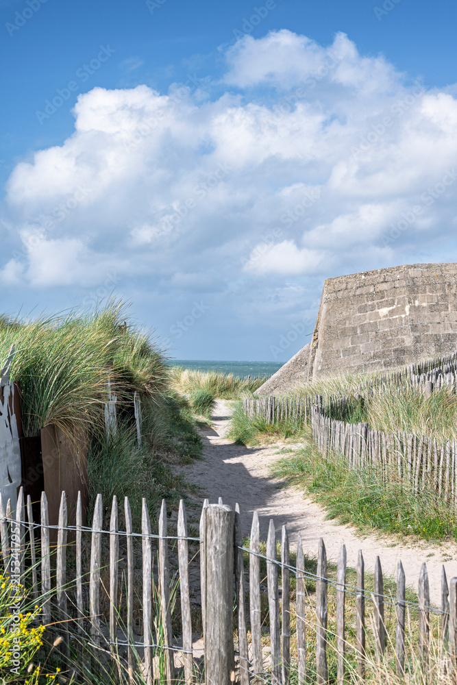 Fototapeta premium A sandy path leading to the sea between wooden fences and an old concrete WWII military bunker on a coastal sand dune at Juno Beach.