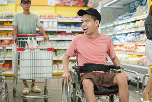 Cheerful young Asian man in wheelchair enjoying grocery shopping in supermarket. Happy disabled shopper living an active independent lifestyle with accessibility, inclusion and diversity concept.