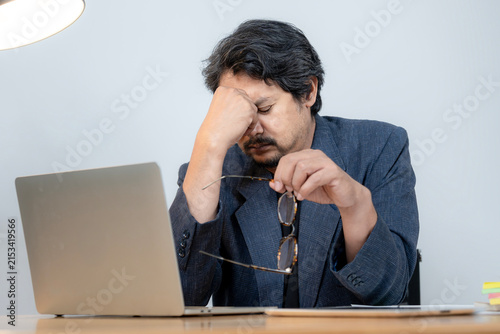 A stressed Asian male office worker sitting at his desk, overwhelmed by work pressure.