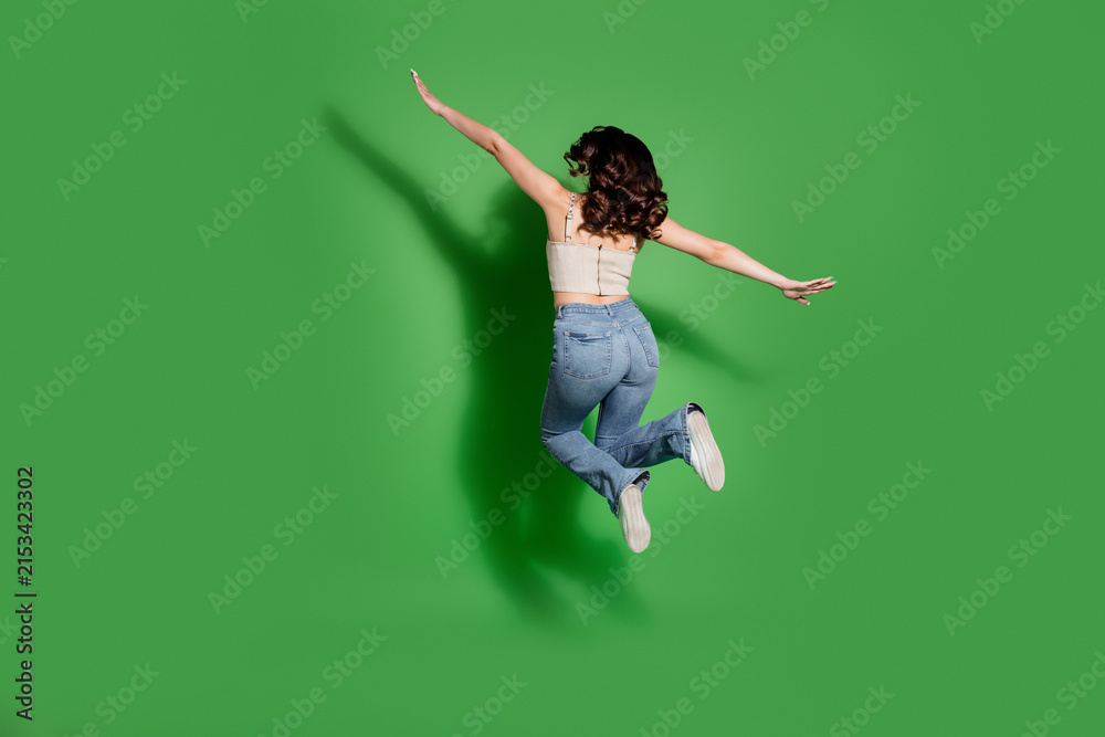 Fototapeta premium Woman jumping midair in jeans on a green background expressing joy, motion and carefree energy in a studio fashion lifestyle pose