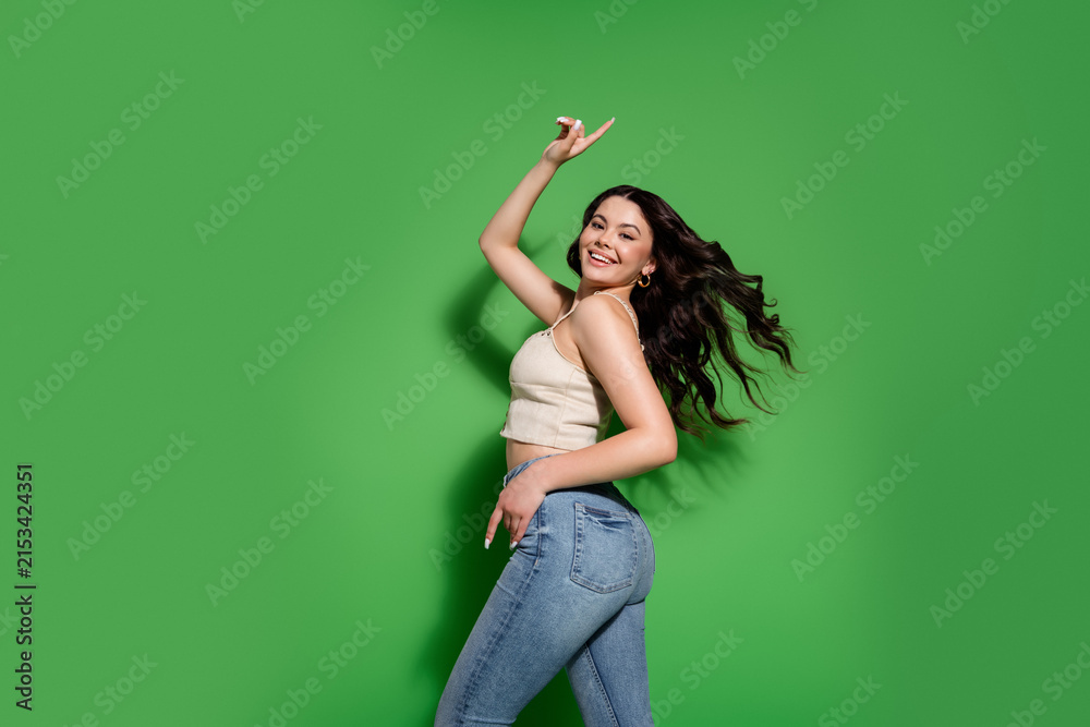 Fototapeta premium Young smiling woman dancing in jeans and a crop top against a bright green background expressing joy and confident casual style