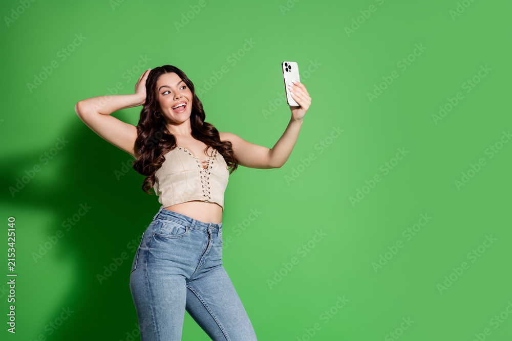 Fototapeta premium Happy young woman taking a selfie with smartphone against green background, casual crop top and jeans, influencer-style portrait showing joy and confidence