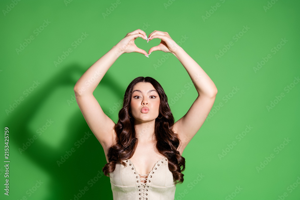 Fototapeta premium Young woman making a heart gesture above her head and blowing a kiss against a green studio background
