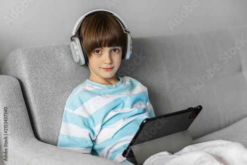 Cute boy in headphones with a digital tablet sitting on a sofa and looking at camera. Kids online education and lifestyle.