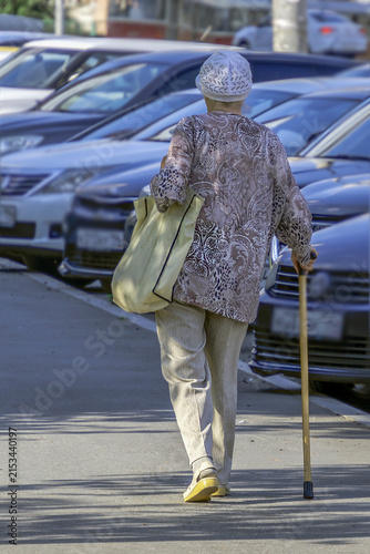 An elderly woman walks along the sidewalk with shopping on a summer day