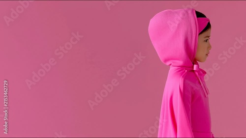 A young girl in a vibrant pink hooded outfit poses against a solid pink background, crossing her arms and turning to the side.