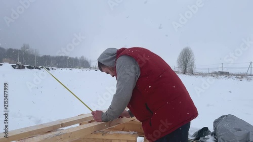 Person in red vest bends over timber structure in snowy rural field with bare trees, utility poles, falling snow, gray hoodie, and overcast winter light during focused outdoor work