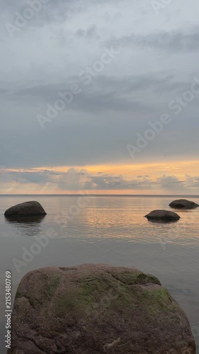 Calm coastal waters beneath broad overcast sky, warm sunset glow along distant horizon, low rocky shoreline, mossy foreground ledge, smooth reflections, soft gray blue orange tones, quiet spacious