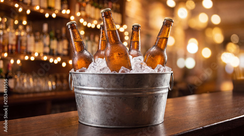 Bottles of beer in a metal bucket filled with ice on a wooden bar counter, illuminated by warm bokeh lights in the background