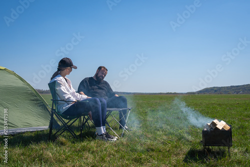 Couple relaxing in camping chairs by a green tent, enjoying a small wood-burning campfire under a clear blue sky in an open grassy field, peaceful summertime escape