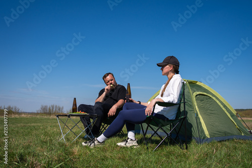 Two people are relaxing in camping chairs next to a tent in a green field under a clear blue sky, enjoying a snack and drinks during their outdoor leisure time
