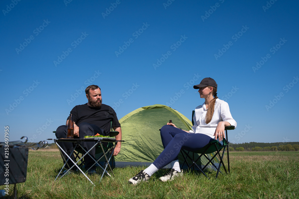 Fototapeta premium Couple resting and enjoying drinks in camping chairs next to their tent in a sunny green field, symbolizing leisure, outdoor activity, and a shared travel experience