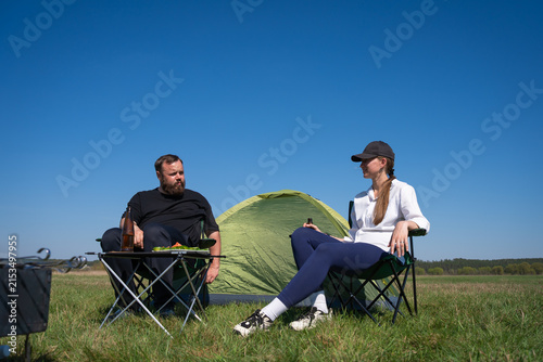 Couple resting and enjoying drinks in camping chairs next to their tent in a sunny green field, symbolizing leisure, outdoor activity, and a shared travel experience