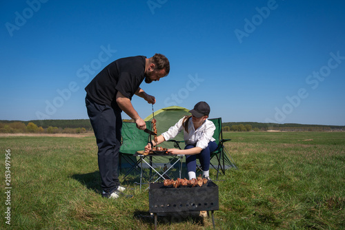 Man and woman preparing grilled meat skewers on a portable barbecue grill, savoring an outdoor picnic experience in a green field during summer leisure time with a camping tent