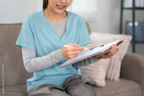 Smiling young Asian healthcare worker writing in patient notebook while sitting on couch in modern medical office