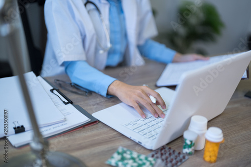 Female doctor in white coat and blue gloves typing on laptop while reviewing patient medication records at clinic desk