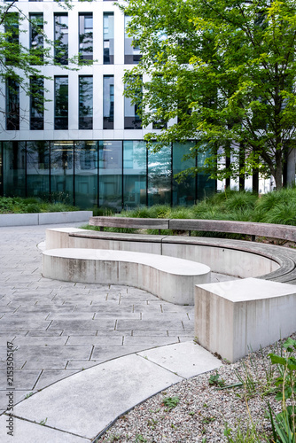 Curved concrete benches trace a courtyard where landscaping and trees soften modern glass architecture, as an urban walkway bends around the planted edge.