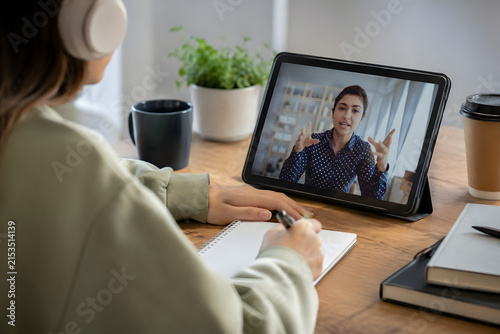 Student taking notes during online video call lecture on tablet with headphones in home workspace.