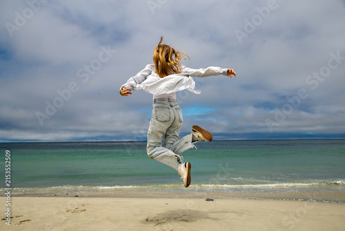 Принти на полотні Person joyfully jumping in mid-air on a sandy beach with the ocean in the background beneath an overcast sky