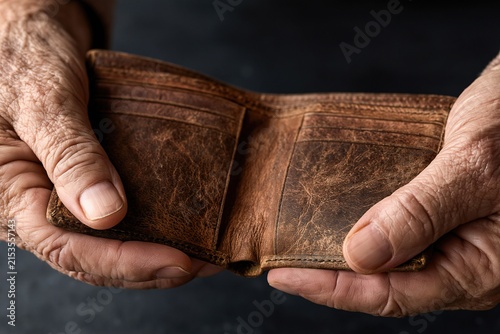 Elderly hands holding an empty brown leather wallet against a dark background symbolizing financial struggle