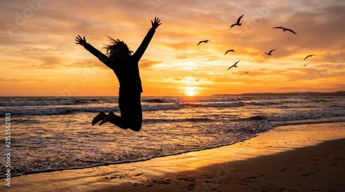 Silhouette of happy woman jumping with arms raised on beach at sunset, freedom concept with seagulls flying over ocean waves during golden hour.
