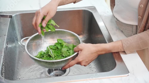 Woman washing fresh green leafy vegetables in metal colander under kitchen sink faucet water