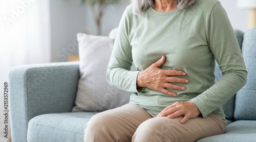 Senior woman stomach pain abdominal discomfort digestive problem sitting couch living room casual clothing hand belly health issue home interior Senior woman with gray hair sitting couch holding