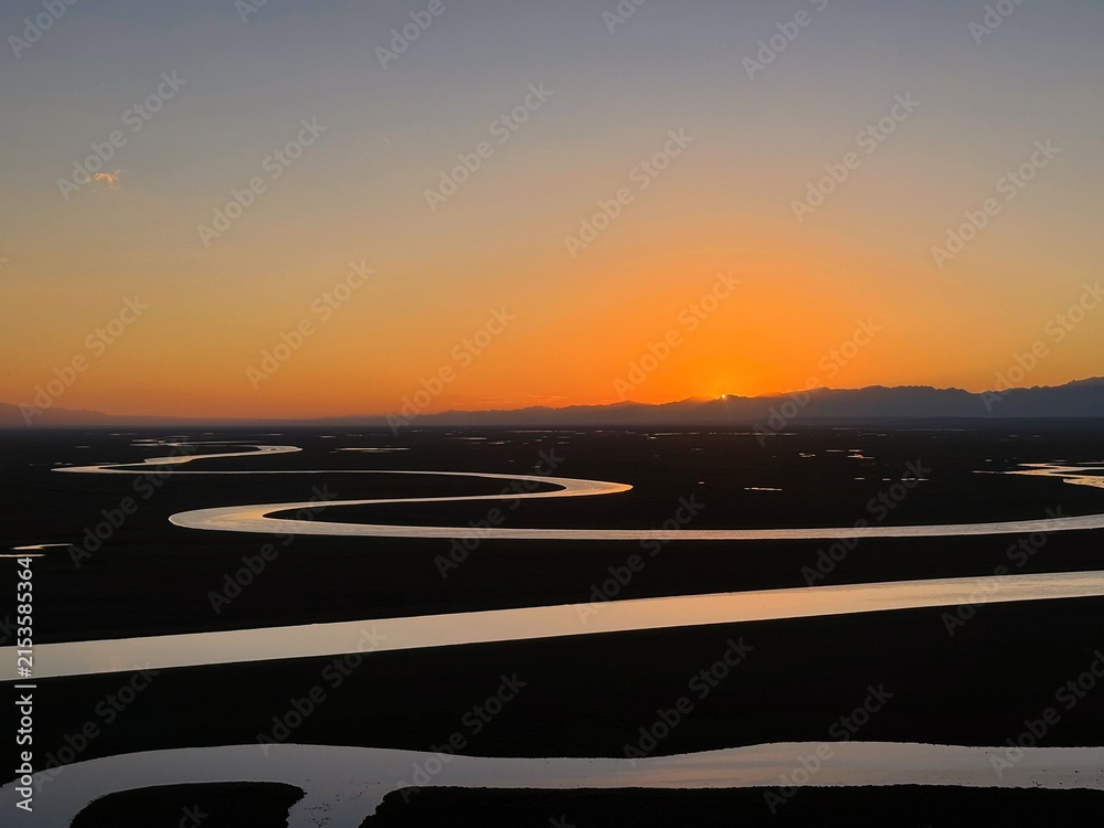 Fototapeta premium A scenic landscape of a winding river reflecting the warm golden light of sunset, with a distant mountain range silhouetted against the evening sky. Peaceful natural scenery with dramatic light and ex