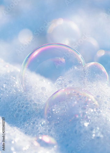 Soap bubbles sit on frothy white foam with soft pastel reflections and a blurred blue background. The composition conveys lightness, cleanliness, and delicate texture.