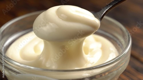 Close-up of a spoon lifting creamy mayonnaise from a glass bowl on wood