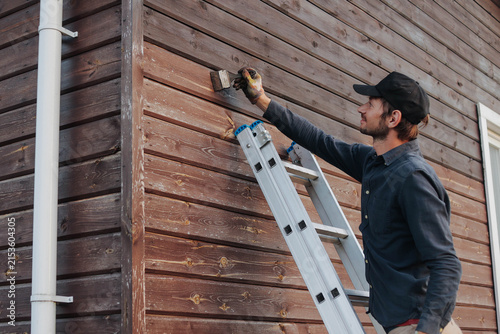 Bild auf Leinwand Professional handyman painting a wooden exterior wall with a brush while standing on an aluminum ladder, home renovation and maintenance concept