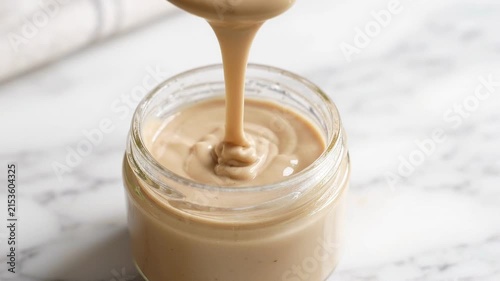 Creamy tahini sauce being poured into a glass jar on a marble surface