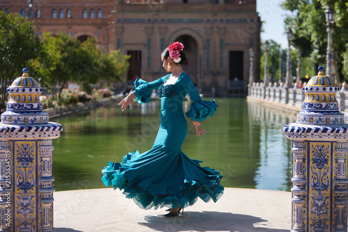 Young, brunette, Andalusian woman, beautiful, wearing a green flamenco dress and flowers in her hair, dancing flamenco, with the canal in the background, in Spain Square in Seville.