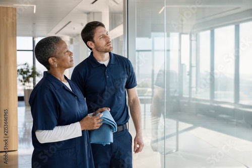 Woman cleaner and male colleague inspecting a freshly cleaned modern office interior. Teamwork, quality control, professional maintenance service, and workplace cleaning concept.