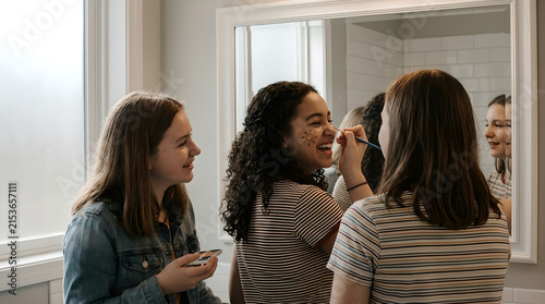 Teenage girls apply makeup together in front of a mirror while preparing for an event.