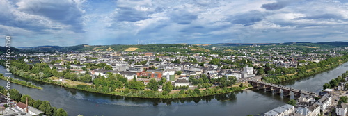Luftbild von Trier mit der antiken Römerbrücke über die Mosel, umgeben von Stadtarchitektur und grünen Hügeln unter bewölktem Himmel an einem Sommertag.
