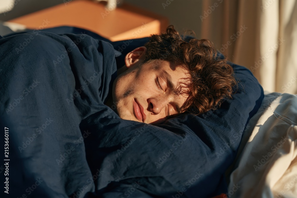 Fototapeta premium Young man wrapped in navy bedding sleeping with sunlight on face for calm bedroom background and wellness banner design